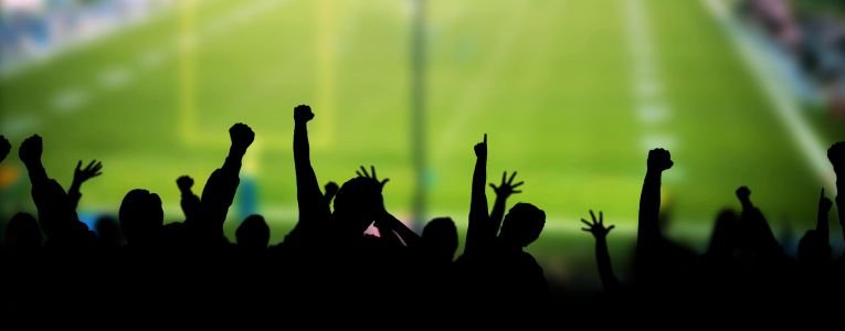 Excited fans of Carey Football in stadium seats cheer for their team, appearing as dark silhouettes against the bright lights of the football field.