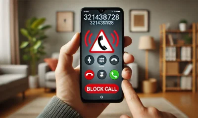 A smartphone displaying an incoming call from the number 3214387828, with a red warning triangle symbol indicating a potential scam. The phone is held in a hand, and the background shows a muted home or office setting, emphasizing caution.